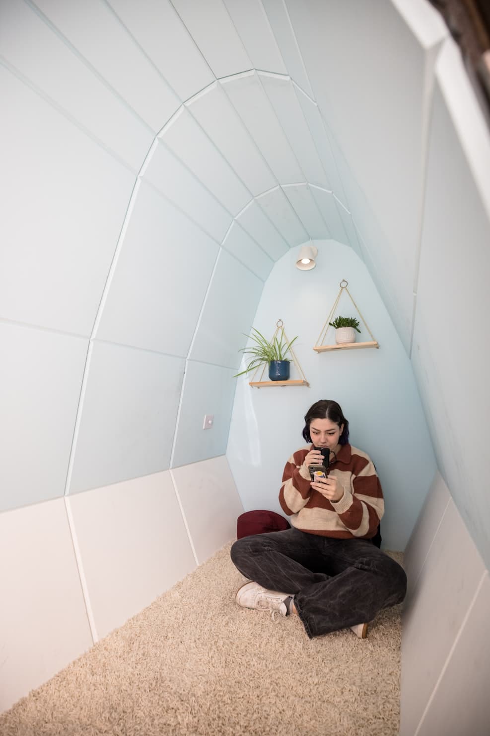 A person sitting cross-legged in a quiet, softly lit pod-like nook, looking calmly at their phone.